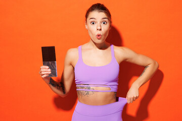 Young fitness trainer instructor sporty woman sportsman wears purple top clothes in home gym hold chocolate bar show loos leggings isolated on plain orange background. Workout sport fit abs concept.