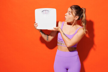 Young shocked fitness trainer instructor sporty woman sportsman wear purple top clothes spend time in home gym hold look at scales isolated on plain orange background. Workout sport fit abs concept.