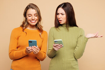 Young confused friends two women they wear orange green shirt casual clothes together hold use show mobile cell phone isolated on plain pastel light beige background studio portrait Lifestyle concept