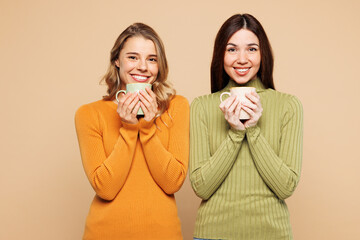Young happy fun friends two women they wear orange green shirt casual clothes together hold cup drink coffee look camera isolated on plain pastel beige background studio portrait. Lifestyle concept.