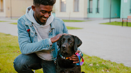 Cheerful african american man spending free day in city with labrador dog. Handsome young guy relaxing with his four-legged cute friend on green lawn.