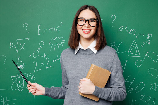 Young smart teacher woman wear grey casual shirt glasses hold in hand book pointer, point on formula isolated on green wall chalk blackboard background studio Education in high school college concept