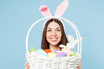 Close up cropped young woman wears pink casual clothes rabbit bunny ears look camera through wicker basket with eggs isolated on plain blue background studio portrait. Lifestyle Happy Easter concept.
