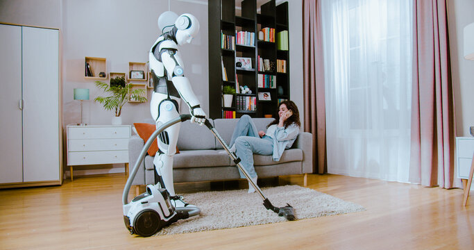 Futuristic cyborg using vacuum cleaner while woman in casual wear lying on comfy sofa and talking on smartphone at home. Concept of robot and human cooperation.