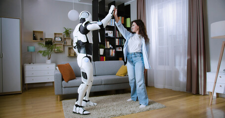 Carefree joyful woman dancing with human like futuristic robot in cozy apartment. Happy caucasian brunette in casual wear having fun and making high five gesture with cyborg.