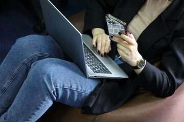 Girl in jeans sitting on padded stool with laptop on her knees and smartphone. Comfortable work in office