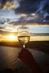 Female hand with glass of white wine against a breathtaking seaside sunset