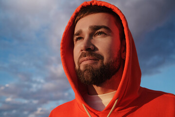 Portrait of a thoughtful man in a vibrant orange hoodie, gazing into the distance under a dramatic...