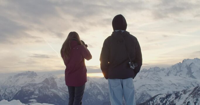 Back shot of Two photographers standing on snow covered mountain capturing sunset