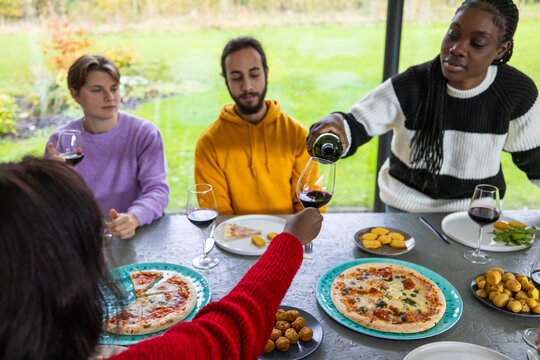 A snapshot of modern diversity, this image depicts a group of multicultural friends of various genders and ages enjoying a convivial meal. They're gathered in a garden-view dining room, sharing