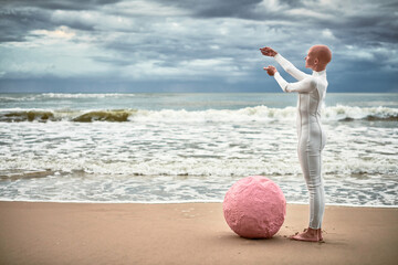 Full length portrait of hairless girl with alopecia in white futuristic suit standing on sea beach...