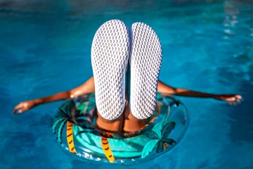 Deurstickers A teenager girl relaxes on a pool ring, her sneaker soles upturned towards the camera, ready for an overlay of text. Leisure, youth, and fun in the sun. © Maria
