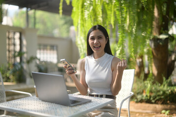 Cheerful young woman celebrating a win on her smartphone at an outdoor garden table with a laptop.