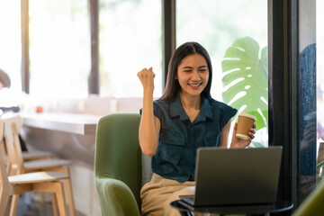 Cheerful young woman celebrates victory with fist pump while working at laptop in sunny cafe.