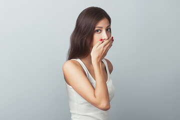 Close-up portrait of young woman in white sleeveless shirt imitating speak no evil concept on gray...