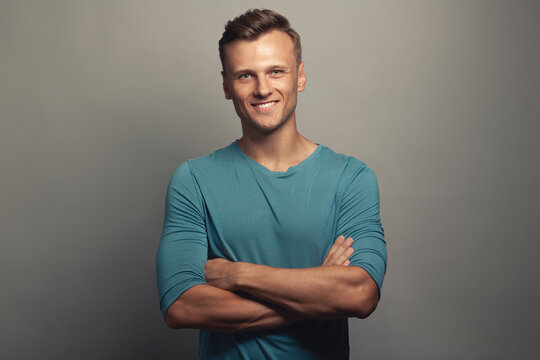 Male beauty concept. Portrait of smiling 30-year-old man standing over gray background with crossed hands. Classic style. Wavy glossy blond hair. Copy-space. Studio shot