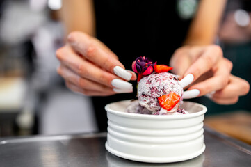 A pair of hands presents a vibrant bowl of ice cream adorned with a flower and fresh fruit—a delightful and appetizing dessert