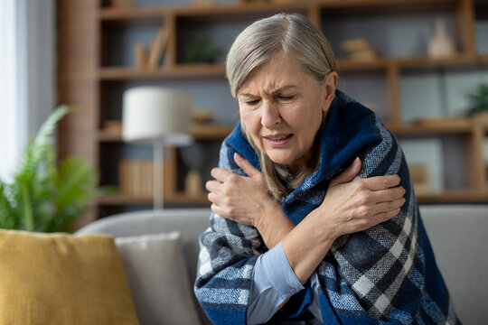 An elderly woman experiencing chills, sitting on the sofa wrapped in a cozy blanket, looking visibly cold and uncomfortable.