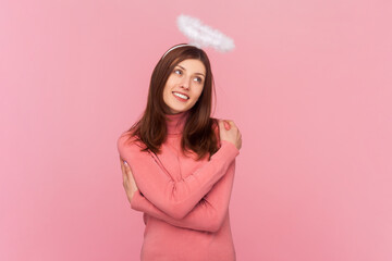 Dreaming woman with brown hair and nimb over head, embracing herself and smiling with pleasure, feeling self-pride, wearing rose turtleneck. Indoor studio shot isolated on pink background