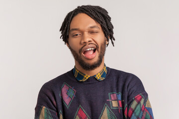 Portrait of playful cheerful attractive african-american man with dreadlocks and beard looking and winking at camera, flirting with somebody. Indoor studio shot isolated on gray background. - obrazy, fototapety, plakaty