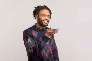 Portrait of smiling satisfied african-american man with dreadlocks and beard standing with mobile phone in hands, recording voice message. Indoor studio shot isolated on gray background.