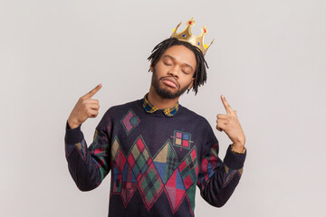 Portrait of proud self-confident handsome african-american man with dreadlocks and beard pointing at golden crown on his head. Indoor studio shot isolated on gray background. - obrazy, fototapety, plakaty
