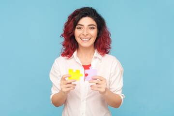 Portrait of smiling beautiful friendly woman with fancy red hair holding piece of puzzles, solving tasks, answering questions, wearing white shirt. Indoor studio shot isolated on blue background.