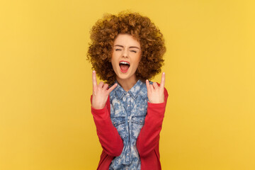 Portrait of excited woman with Afro hairstyle depicting heavy metal rock sign, horns up gesture, screaming with widely open mouth. Indoor studio shot isolated on yellow background. - obrazy, fototapety, plakaty