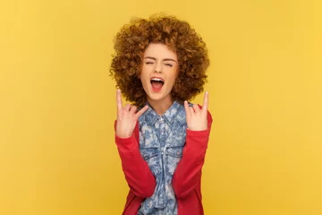 Fotobehang Portrait of excited woman with Afro hairstyle depicting heavy metal rock sign, horns up gesture, screaming with widely open mouth. Indoor studio shot isolated on yellow background. © khosrork