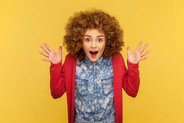 Portrait of emotive woman with Afro hairstyle raises hands and has happy face expression feels excited, being full of happiness. Indoor studio shot isolated on yellow background. - obrazy, fototapety, plakaty