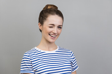 Portrait of cheerful woman wearing striped T-shirt standing and winking playfully, having positive expression, looking at camera. Indoor studio shot isolated on gray background. - obrazy, fototapety, plakaty