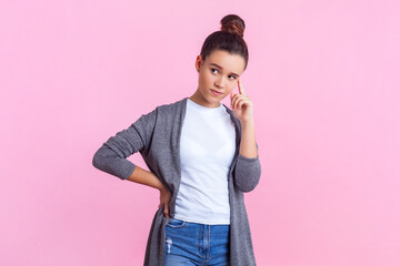 Portrait of concentrated pensive teenage girl with bun hairstyle in casual clothes standing keeps...