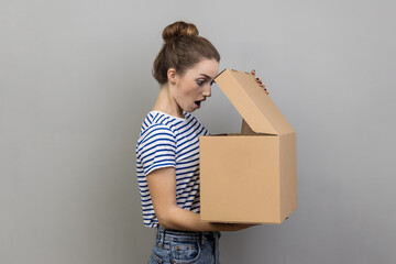 Side view portrait of surprised shocked woman in striped T-shirt standing with carton parcel and...