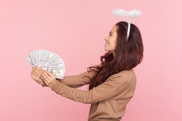 Side view portrait of attractive friendly woman with wavy hair and nimb over head, holding big fan of dollar banknotes, wearing wearing brown pullover. Indoor studio shot isolated on pink background