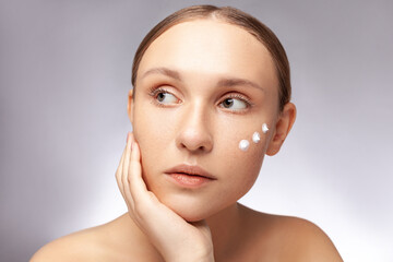 Closeup portrait of beautiful woman with bare shoulders applying creme on cheek looking away with...