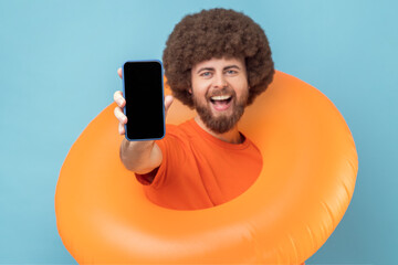 Excited extremely happy man with Afro hairstyle standing with rubber ring and showing smart phone,...