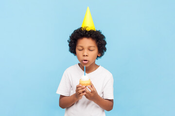 Portrait of cute little boy with curly hair wearing yellow party cone holding cake blowing candle...