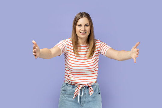 Come into my arms, free hugs. Portrait of adorable hospitable blond woman wearing striped T-shirt smiling and reaching out hands, going to embrace. Indoor studio shot isolated on purple background.