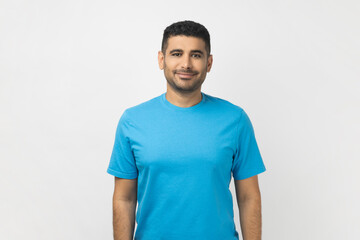 Portrait of happy smiling satisfied unshaven man wearing blue T- shirt standing looking at camera, expressing positive emotions. Indoor studio shot isolated on gray background.