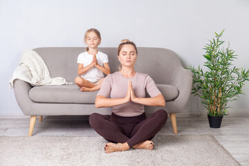 Mindfulness and harmony. Lotus posture. Calm woman with her daughter with closed eyes and prayer gesture practicing yoga together, doing exercise breath technique, meditating in room sitting on floor.