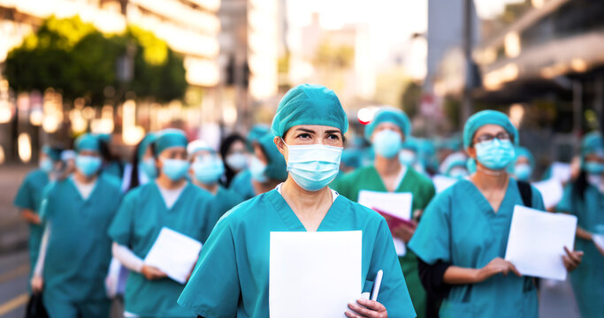 A woman, standing at the forefront holding a blank placard for a message or a demand. Healthcare professionals in medical blue lab coats and masks demonstrate in the street. Concept: change, rights.