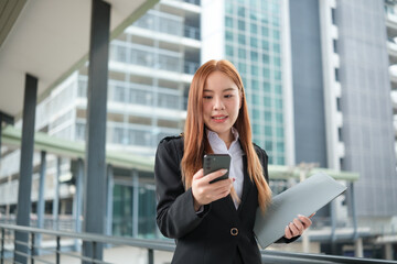 A young Asian businesswomen wearing a suit holding files standing in a big city on a busy downtown street. Young Asian businesswoman using smartphone texting to contact clients.