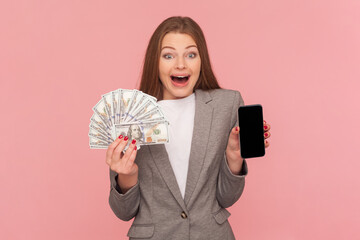 Portrait of amazed surpised woman with brown hair holding dollar banknotes and smart phone with empty display, wearing business suit. Indoor studio shot isolated on pink background.