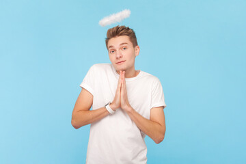 Portrait of cute angelic young man wearing white t-shirt and with nimb over head, pleading with praying gesture, looking at camera. Indoor studio shot isolated on blue background