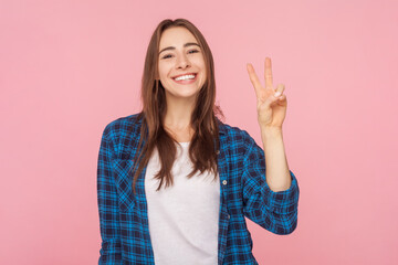 Portrait of smiling cheerful attractive brown haired woman showing peace v sign victory gesture, wearing checkered shirt. Indoor studio shot isolated on pink background.