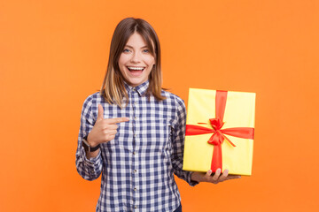 Portrait of cute smiling amazed woman with brown hair pointing at present box celebrating holiday congratulating, wearing checkered shirt. Indoor studio shot isolated on orange background