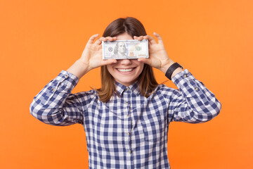 Portrait of happy shy rich woman with brown hair covering her eyes with dollar banknotes smiling to camera, wearing checkered shirt. Indoor studio shot isolated on orange background