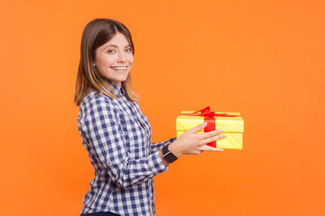 Profile portrait of smiling happy optimistic woman with brown hair giving present to boyfriend looking at camera, wearing checkered shirt. Indoor studio shot isolated on orange background