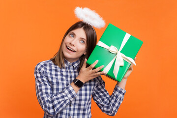 Portrait of cute curious woman with brown hair and nimb over head shaking present box thinking what inside, wearing checkered shirt. Indoor studio shot isolated on orange background