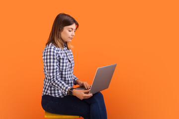 Side view portrait of confident woman with brown hair typing on laptop keyboard, working remotely, wearing checkered shirt. Indoor studio shot isolated on orange background